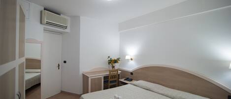 A hotel room with a bed, a desk, a chair, an air conditioner, a nightstand, and a wall-mounted lamp.