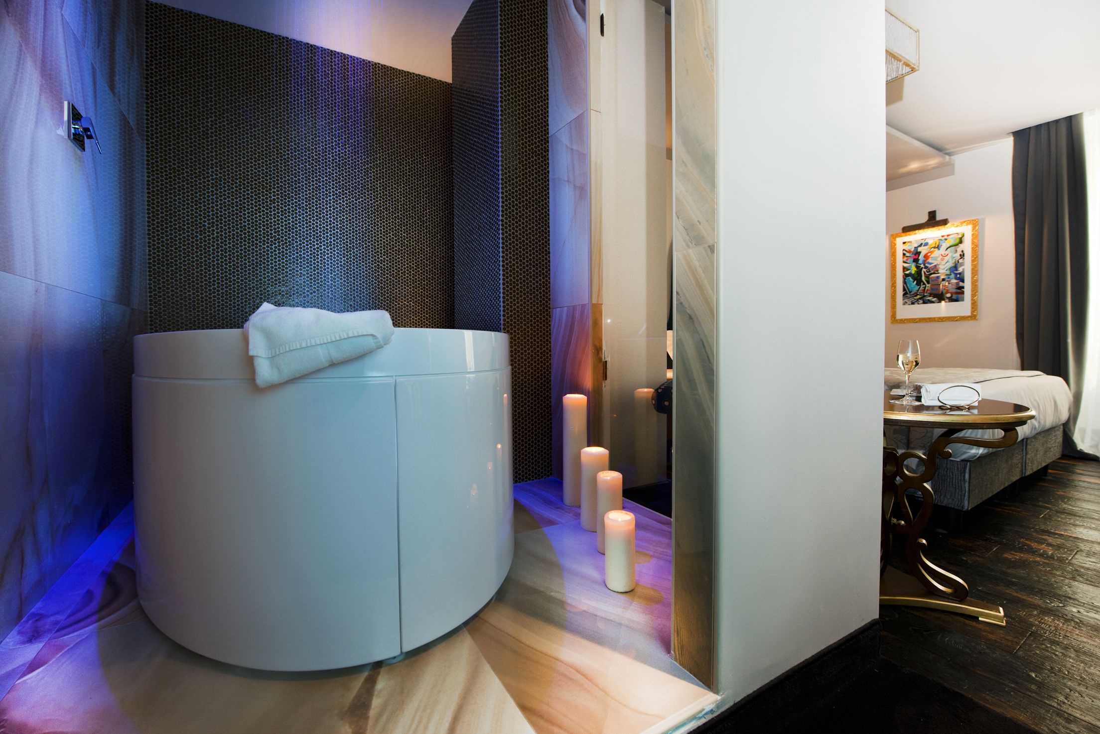 A modern bathroom with a large bathtub, candles, and a bed in the adjacent room.