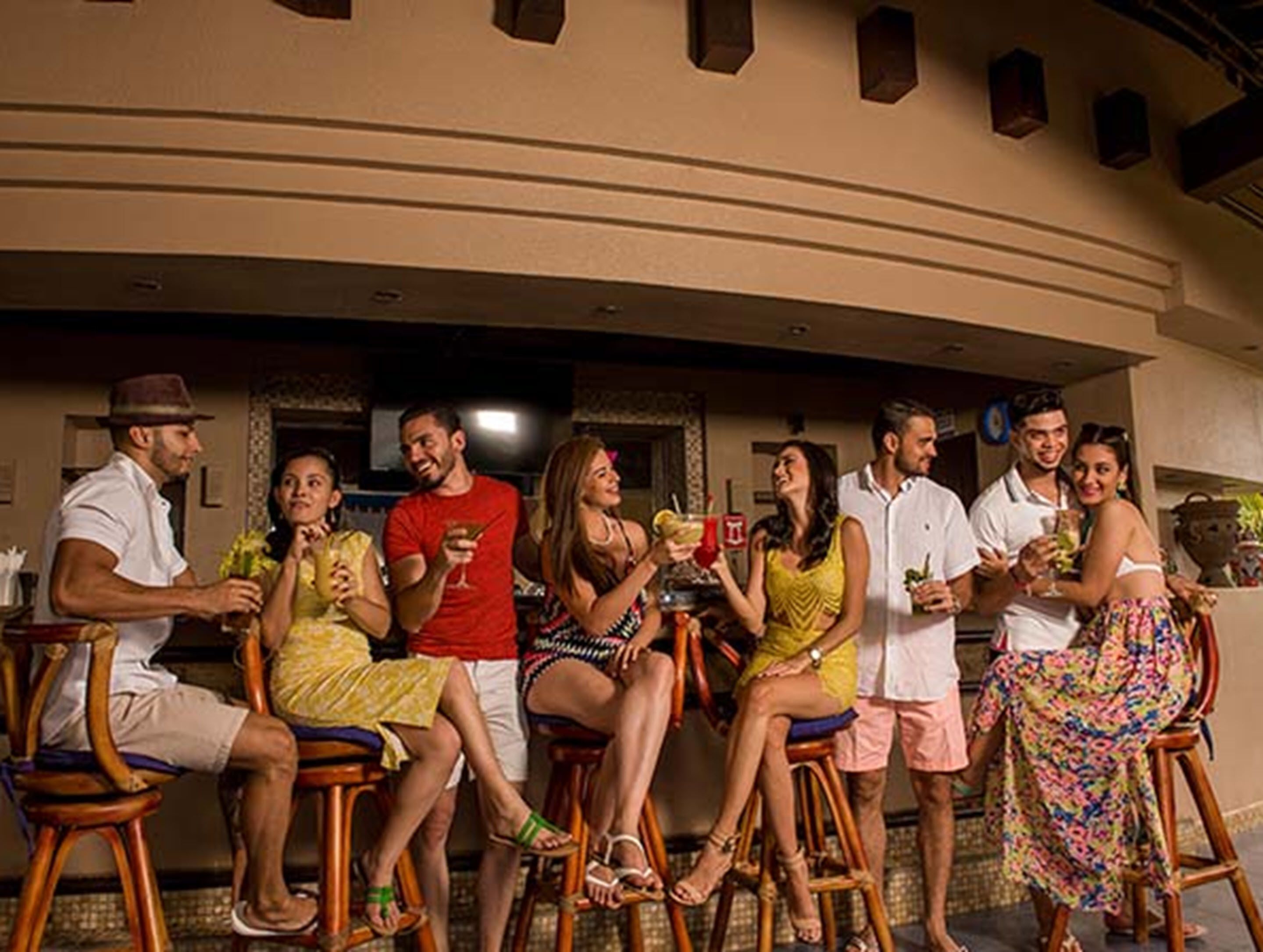 A group of people seated at a bar area with stools, a television screen, and a wall-mounted shelf with items.