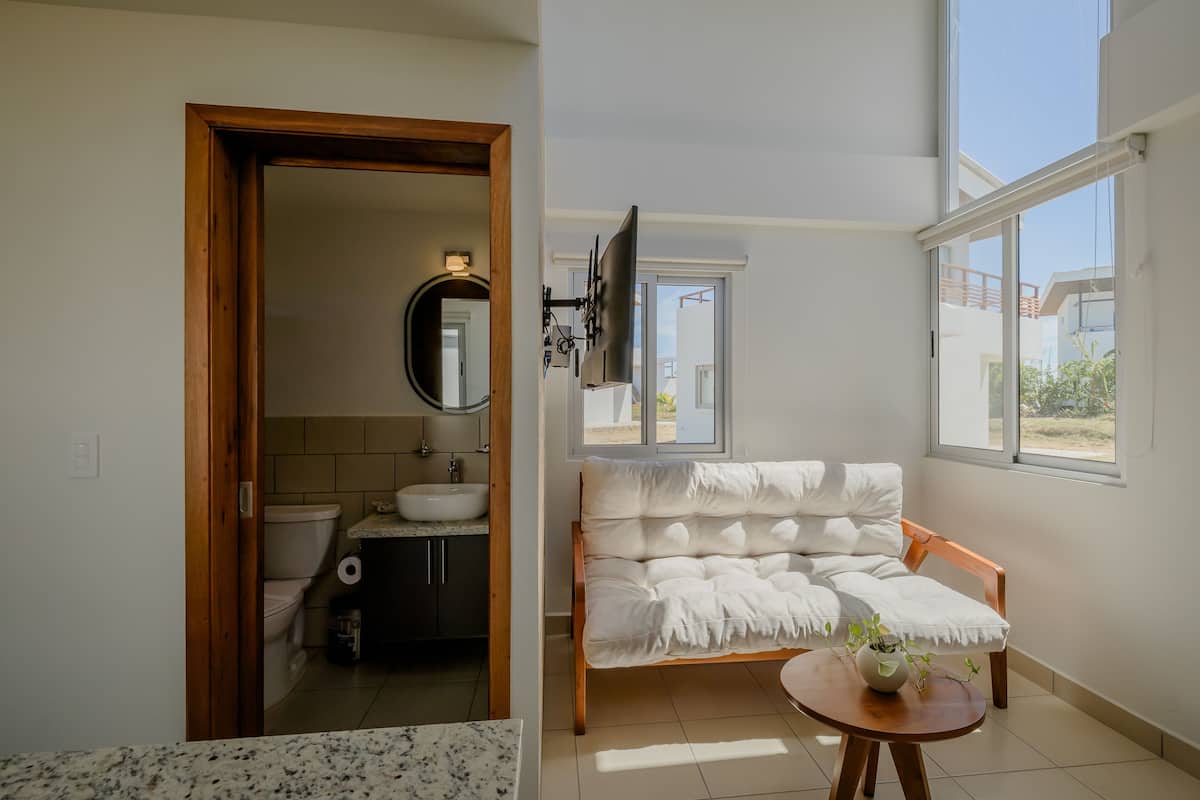 Ein modernes Badezimmer mit Toilette, Waschbecken und Spiegel. Eine Holztür führt in einen anderen Raum, und ein Fenster sorgt für natürliches Licht.