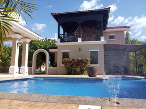 A swimming pool with a water fountain, a covered outdoor area, and a view of a residential building. - Gran Pacifica Beach Resort & Homes (Villa El Carmen)
