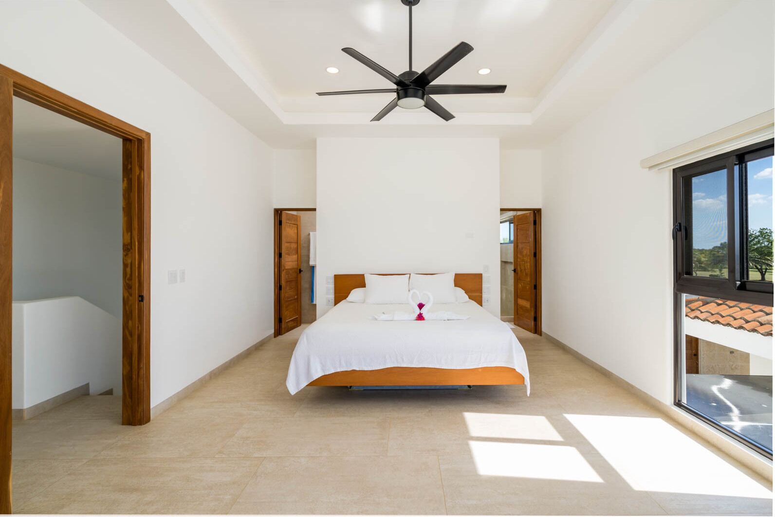 A spacious bedroom with a large bed, a ceiling fan, and a view of the outdoors through a large window.