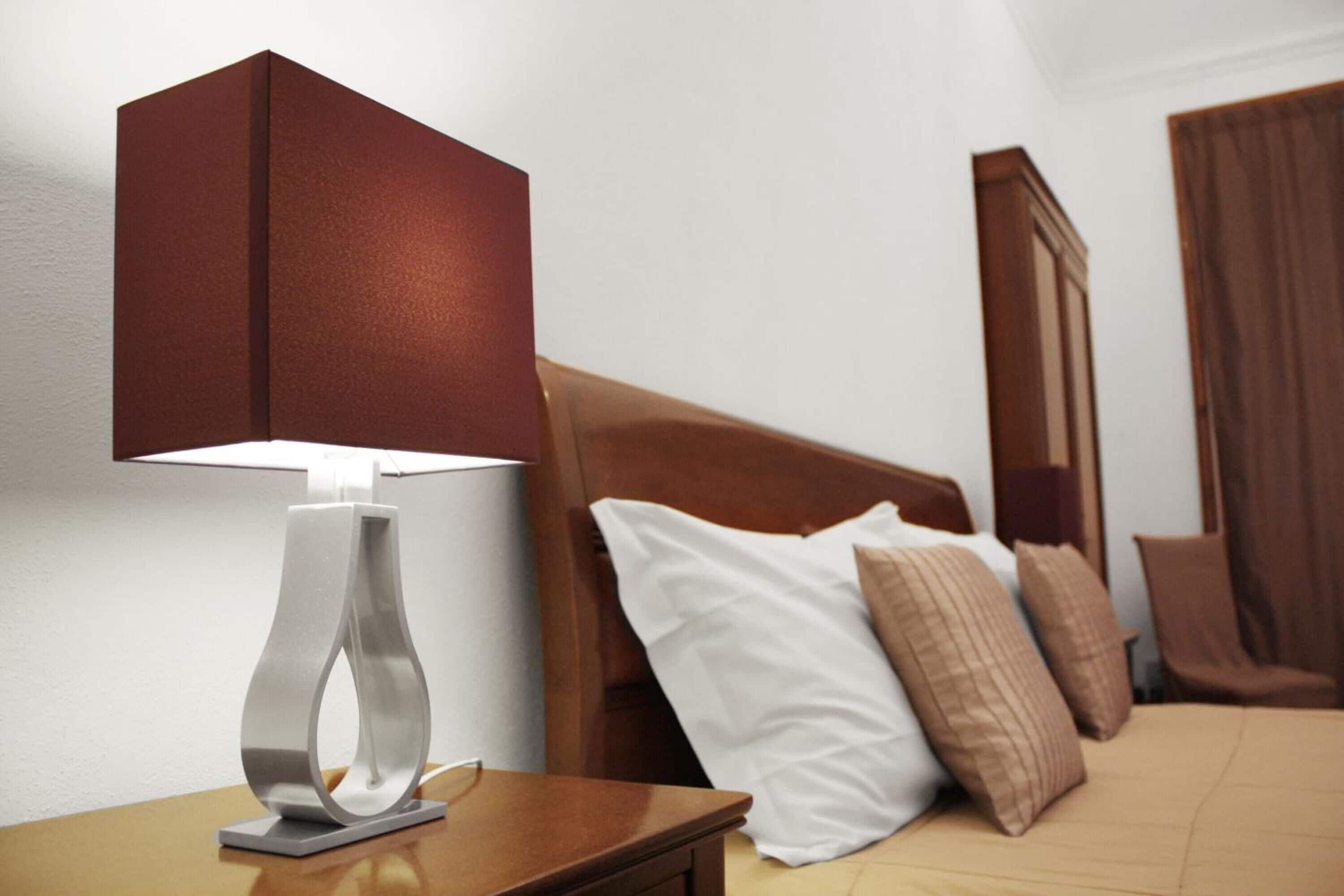 Uma cama bem arrumada com cabeceira de madeira, uma mesa de cabeceira com abajur moderno e uma cadeira de madeira em um quarto de hotel.