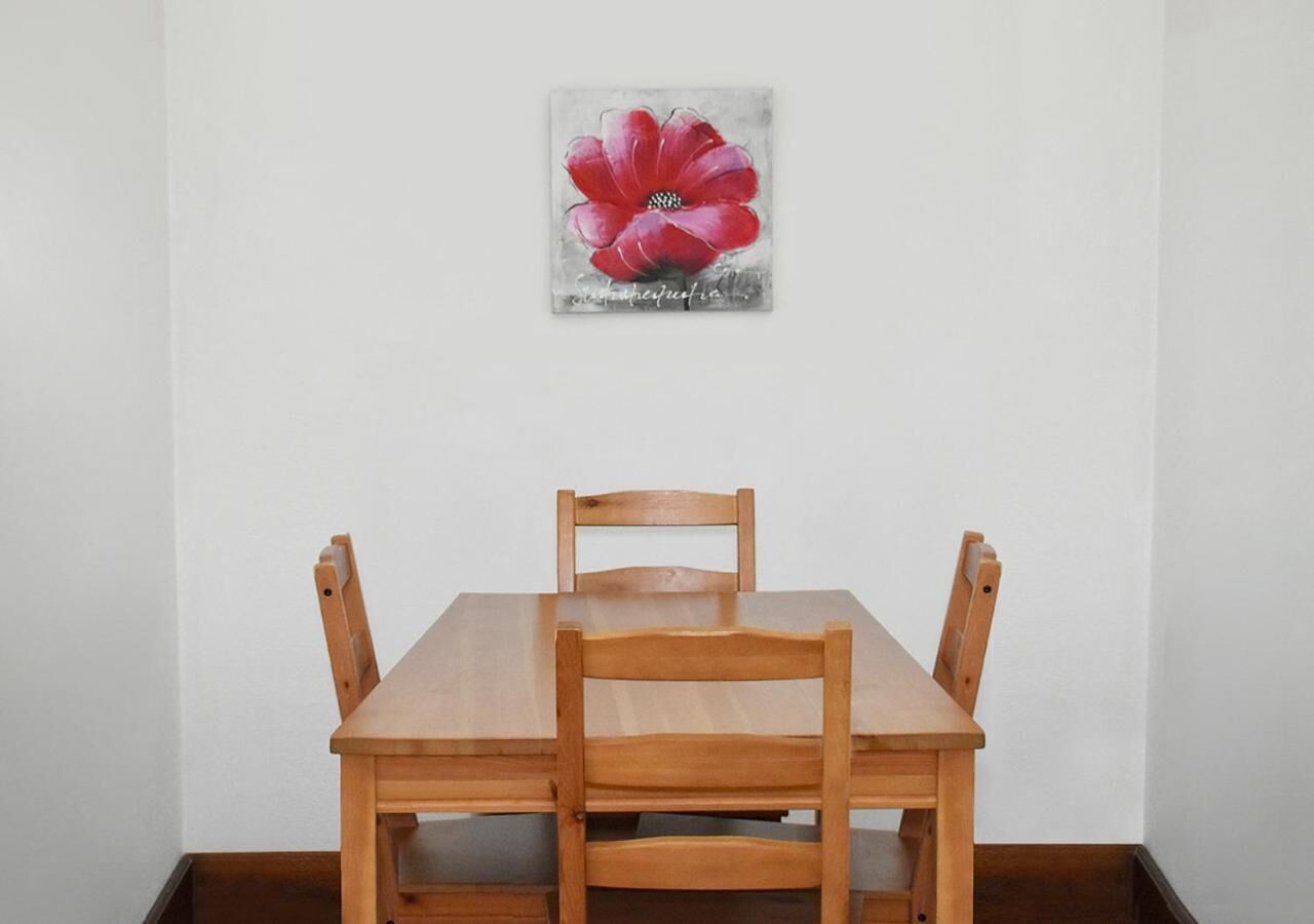 A wooden dining table with four chairs, a painting of a red flower on the wall, and a white wall.