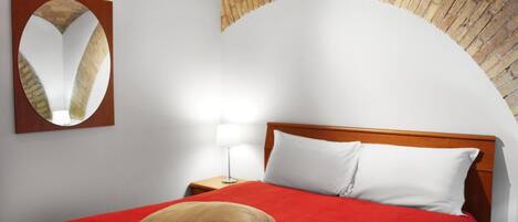 Habitación de hotel con colcha roja, dos almohadas, cabecera de madera, mesita de noche con lámpara y espejo en la pared.