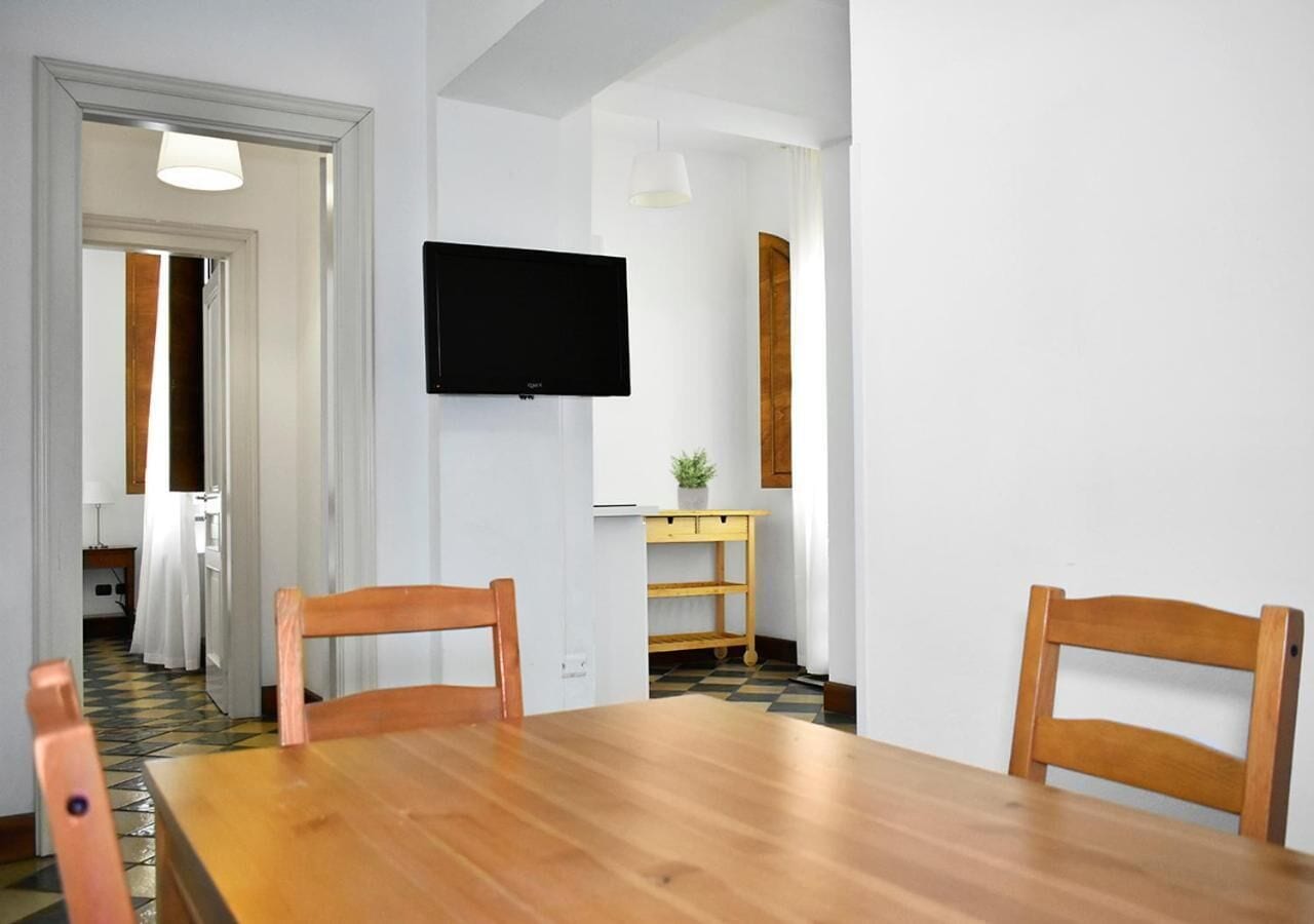 A dining area with a wooden table and chairs, a TV mounted on the wall, and a small wooden side table with a plant.