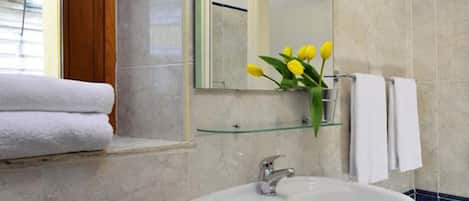 Un baño con un lavamanos blanco, un espejo y un florero con flores amarillas. Hay toallas colgadas en la pared y una ventana con vistas al exterior.