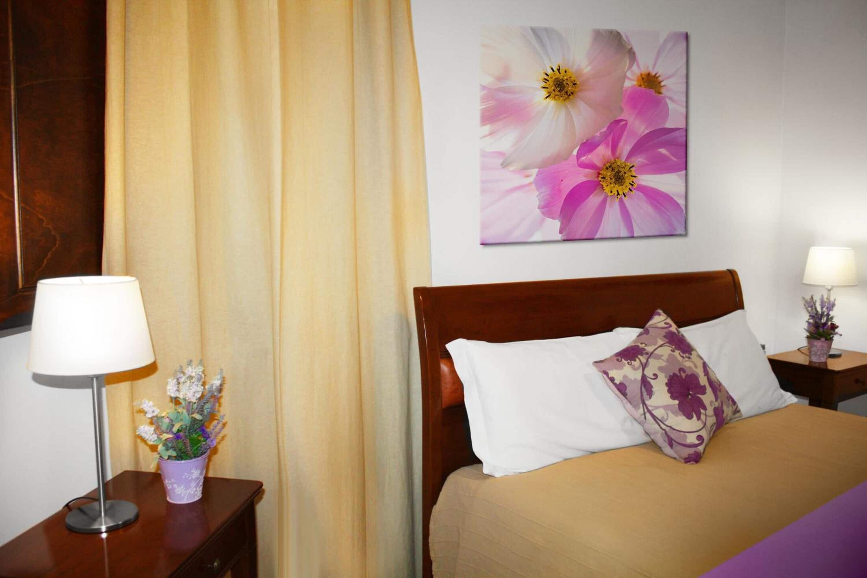 Habitación de hotel con cama, mesita de noche con lámpara, un cuadro de flores en la pared y una cortina.