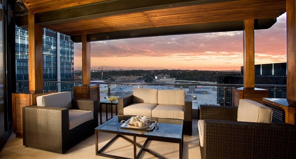 Un salón en la azotea con mobiliario de mimbre, una mesa de centro de vidrio y vistas a la ciudad al atardecer.