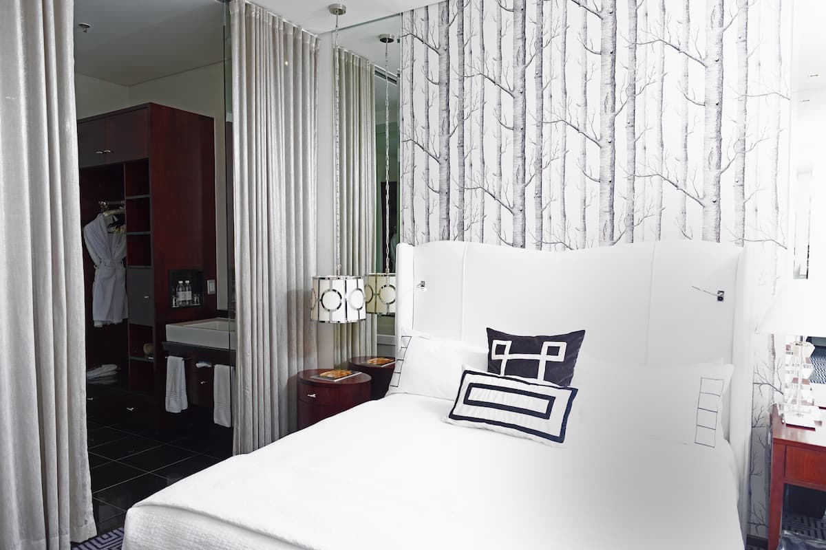 Een hotelkamer met een bed, nachtkastje, spiegel, kledingkast en een badkamer zichtbaar door een glazen deur.