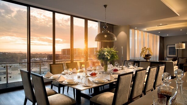 Comedor con mesa preparada para seis personas, ventanas amplias con vista a la ciudad y una lámpara colgante.