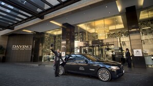 Un coche de lujo negro estacionado frente a la entrada del Hotel y Suites Davinci, con fachada de vidrio y una gran lámpara de araña.