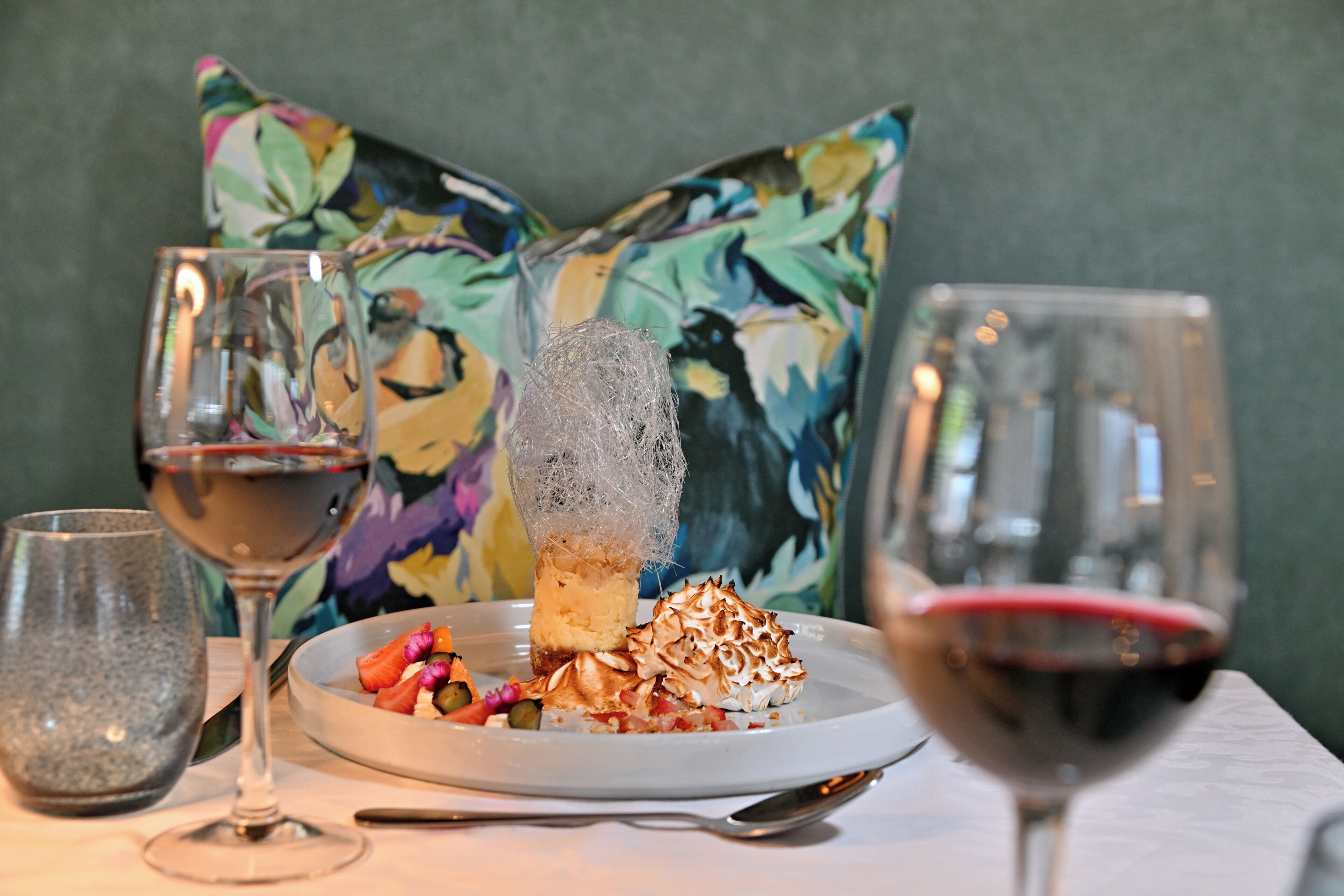 A dining table with a plate of food, two wine glasses, and a floral pillow.