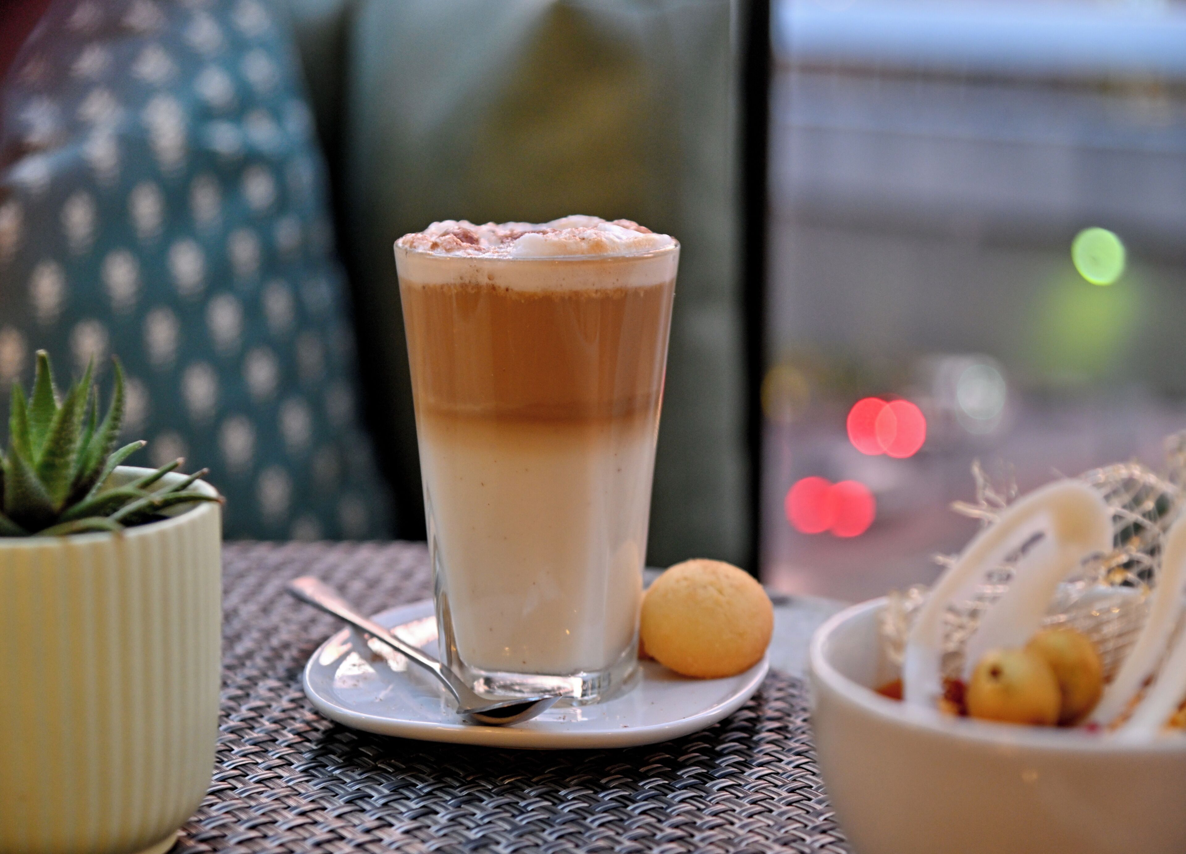 A tall glass of layered beverage on a table with a spoon, a small potted plant, and a bowl of fruit.