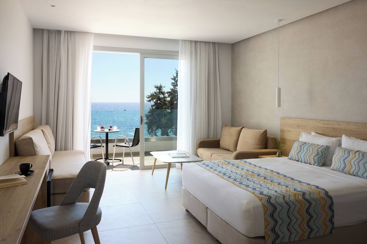 A hotel room with a large bed, a desk, a chair, a small table, and a balcony with a view of the sea.