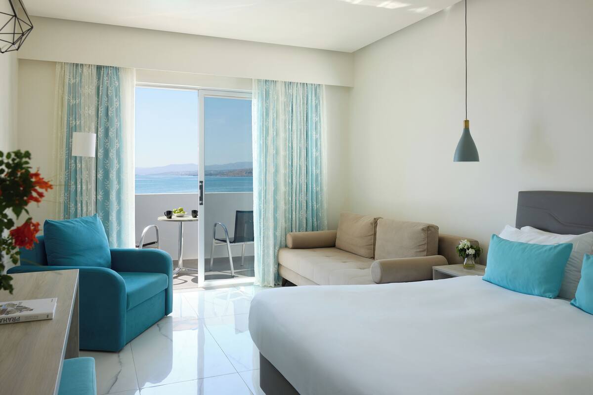 A modern hotel room with a large bed, two sofas, a dining table, and a balcony with a view of the sea.