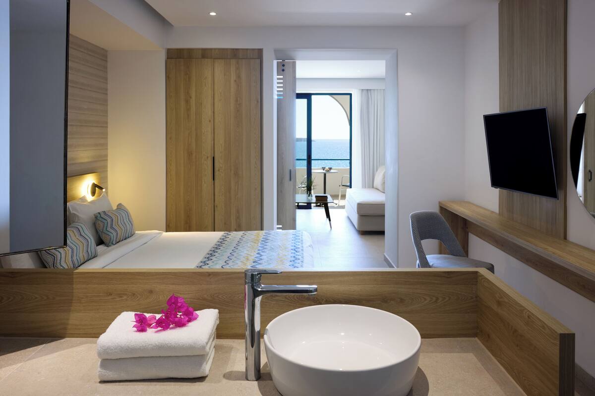 A modern hotel room with a large bed, a wooden vanity with a bowl sink, a flat-screen TV, and a balcony with a view of the sea.