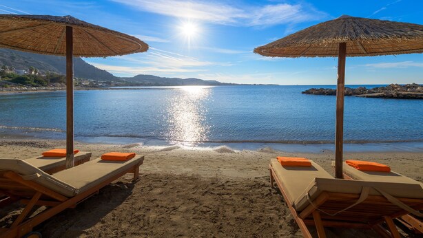 A beach with two wooden lounge chairs under thatched umbrellas, facing a calm sea and a clear sky. - Sunrise Hotel (Rhodes)