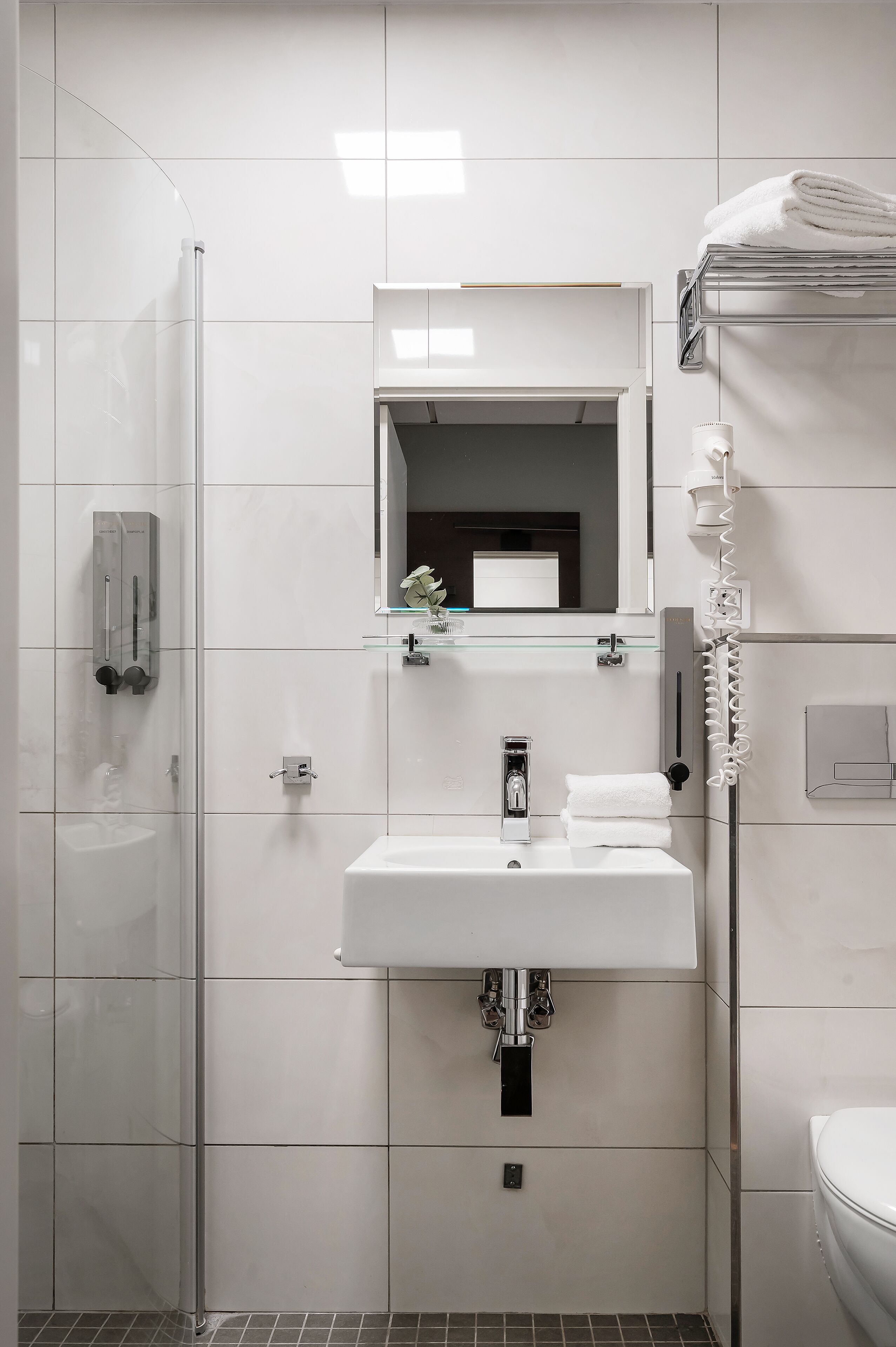 Une salle de bain moderne avec une douche à l’italienne en verre, un lavabo blanc avec un miroir au-dessus et des toilettes avec un porte-serviettes.