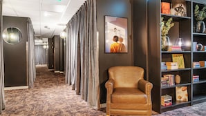 Un hall de hotel con una butaca de cuero marrón, una estantería con libros y objetos decorativos, un cuadro en la pared y una alfombra estampada.