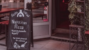 Entrada a un restaurante con un cartel que anuncia NapoliLitan Pizza, mostrando opciones de menú como pasta, ensalada y alternativas veganas.