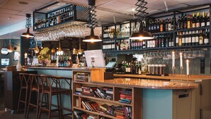 Zona de bar con estantes llenos de botellas, vasos y un monitor de computadora. Hay sillas de madera y una estantería con libros.
