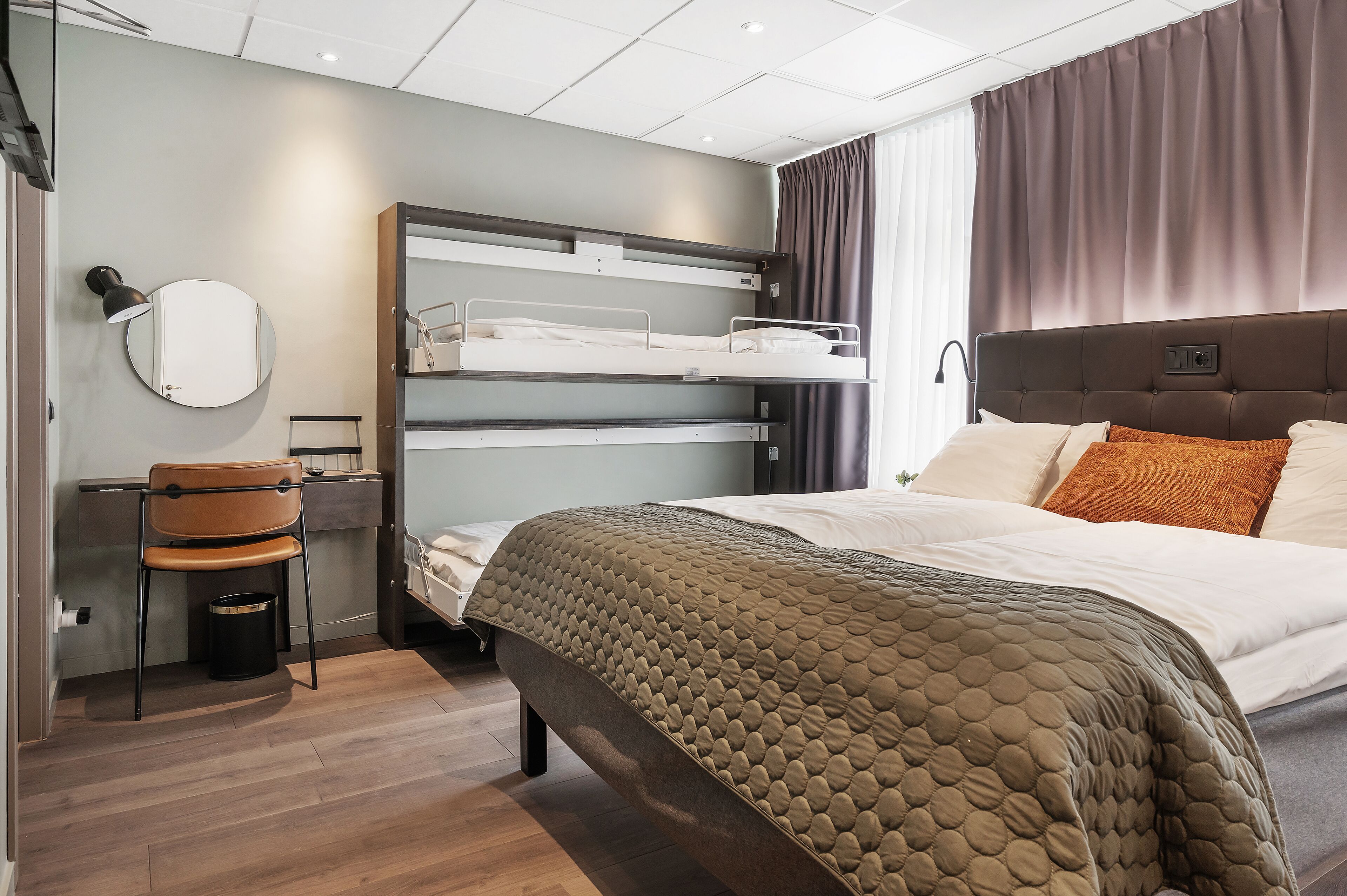 Ett modernt hotellrum med en våningssäng, ett skrivbord, en stol och en stor säng med ett quiltat överdrag.