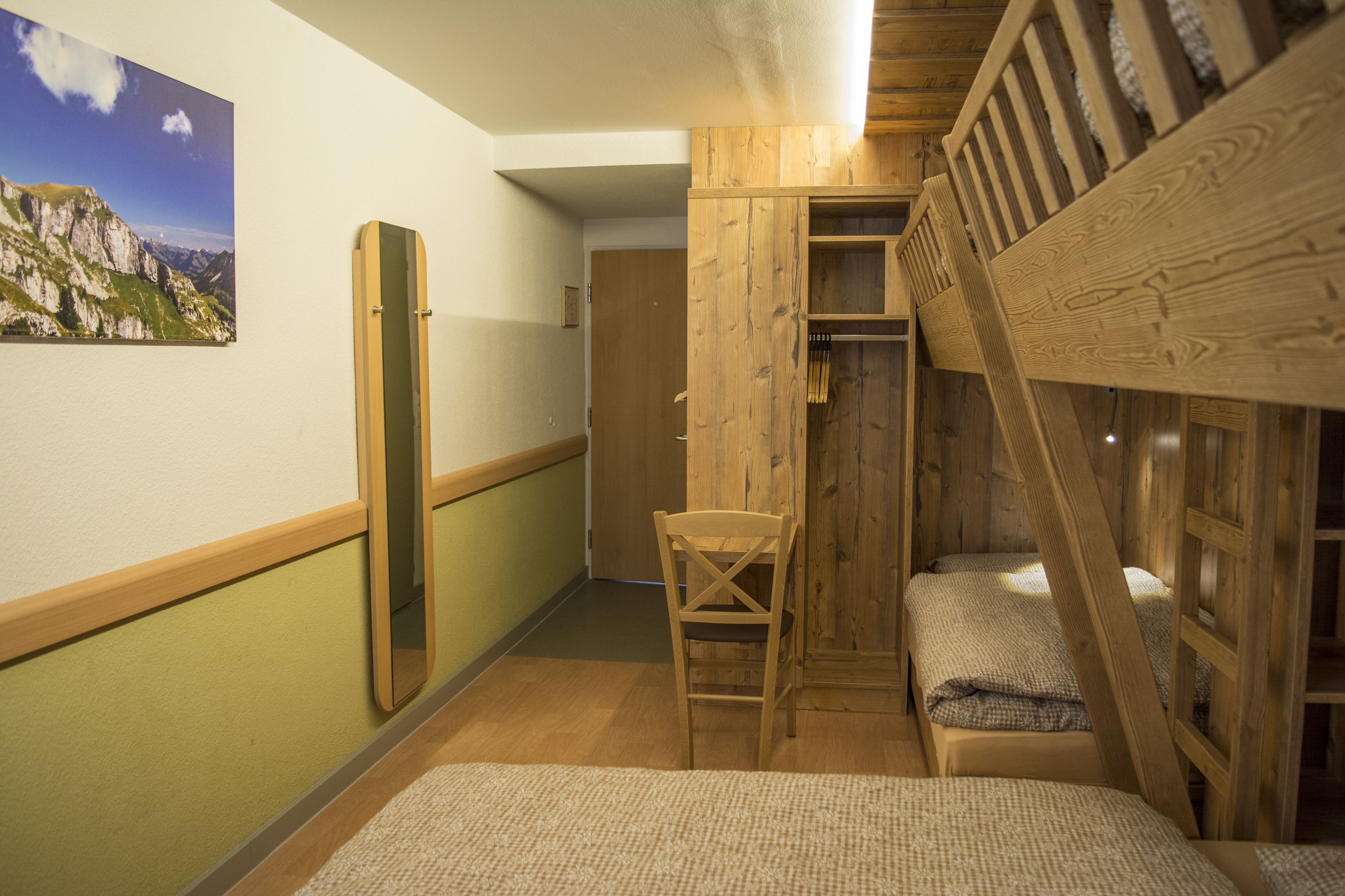 A room with a bunk bed, a wooden wardrobe, a chair, and a mirror on the wall.