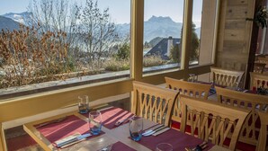 Une table à manger dressée avec des couverts et des verres, des chaises en bois et une vue sur les montagnes par de grandes fenêtres.