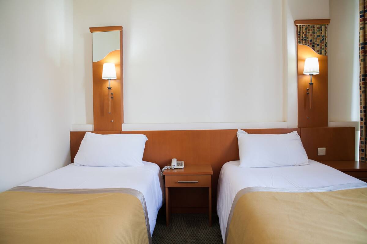 A hotel room with two single beds, a bedside table with a lamp, a phone, and a mirror on the wall.