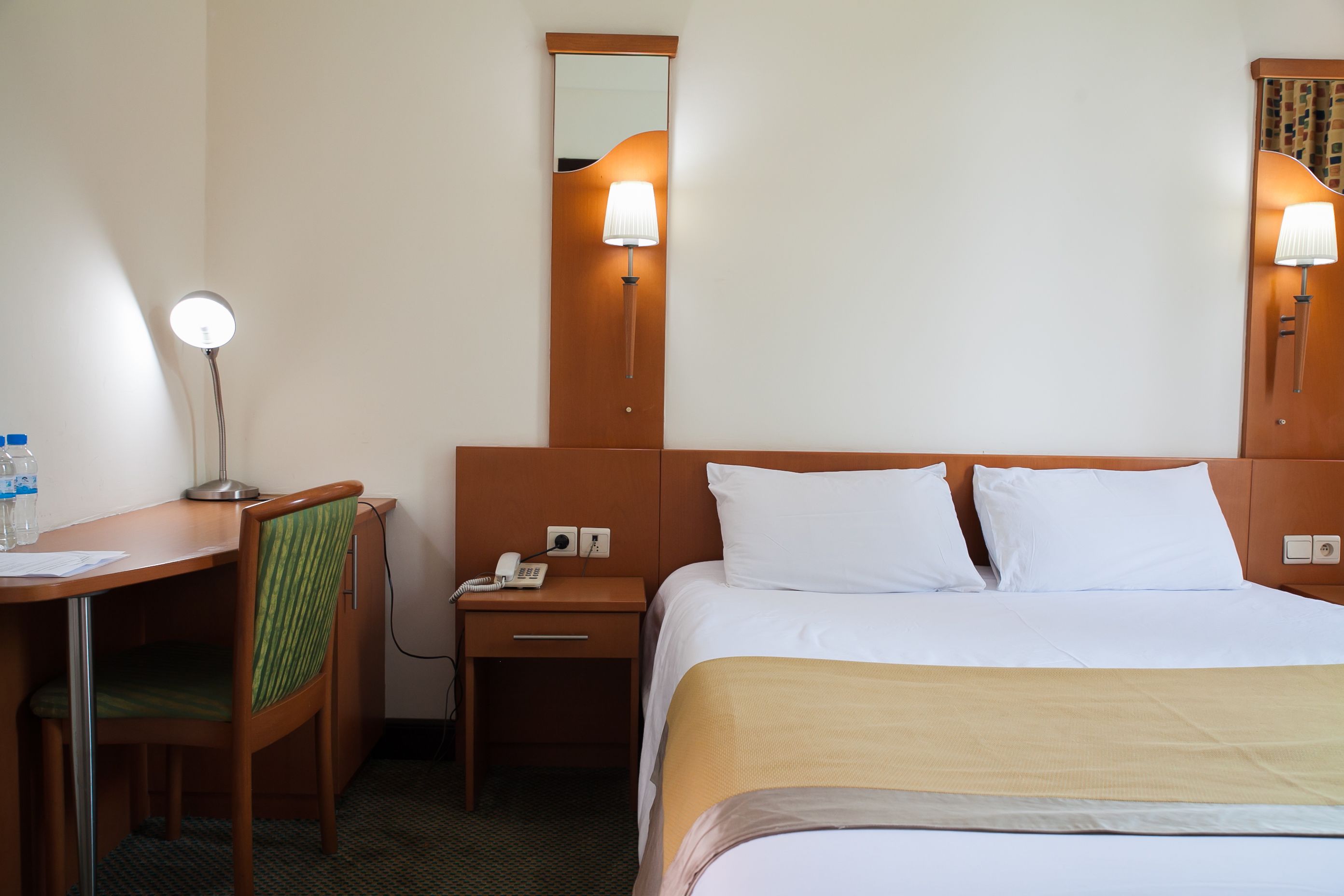 A hotel room with a bed, a desk, a chair, a nightstand, a lamp, and a mirror.
