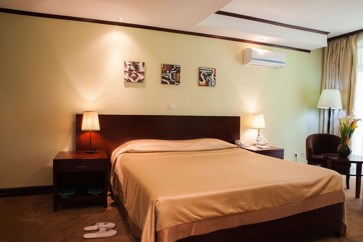 A hotel room with a large bed, two bedside tables with lamps, a chair, a small table with a vase, and artwork on the wall.
