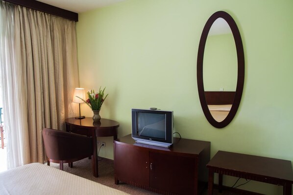 Habitación de hotel con cama, escritorio con televisor, silla, mesita, lámpara y espejo en la pared.