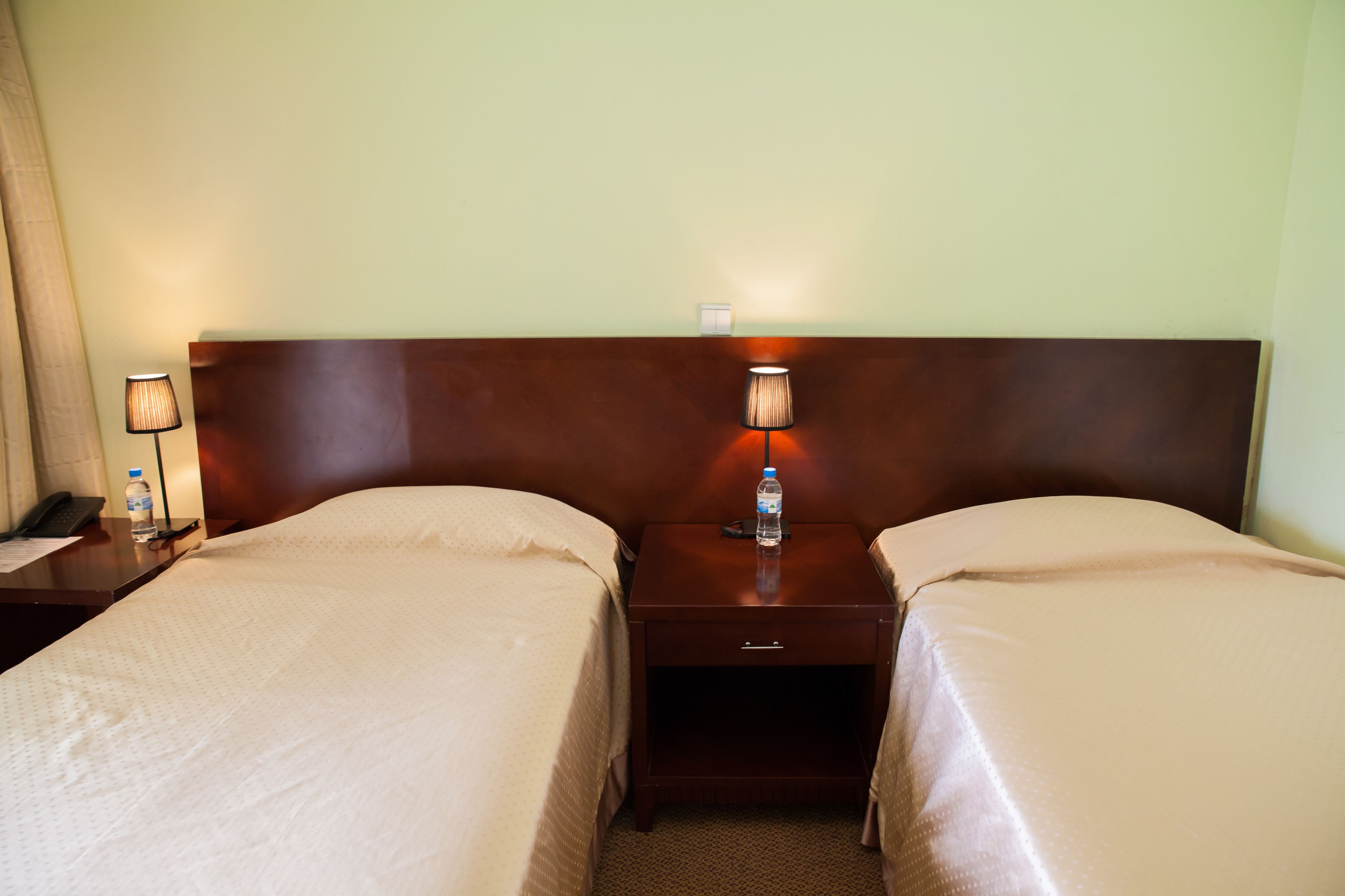 A hotel room with two single beds, a wooden headboard, bedside lamps, a nightstand with a phone and a water bottle, and a carpeted floor.