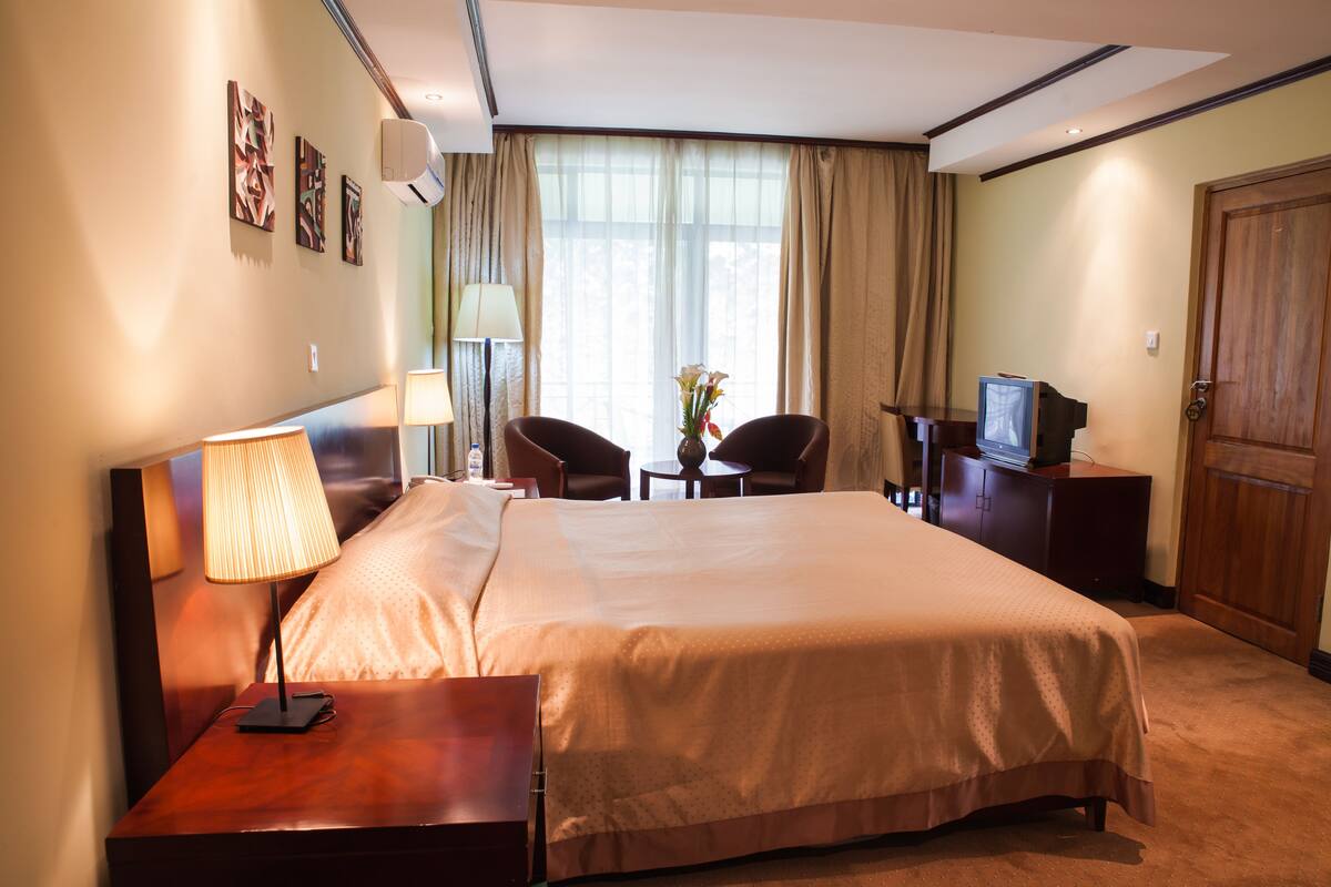Une chambre d’hôtel avec un grand lit, des tables de chevet, un bureau avec une lampe, deux chaises, une télévision et une fenêtre avec des rideaux.
