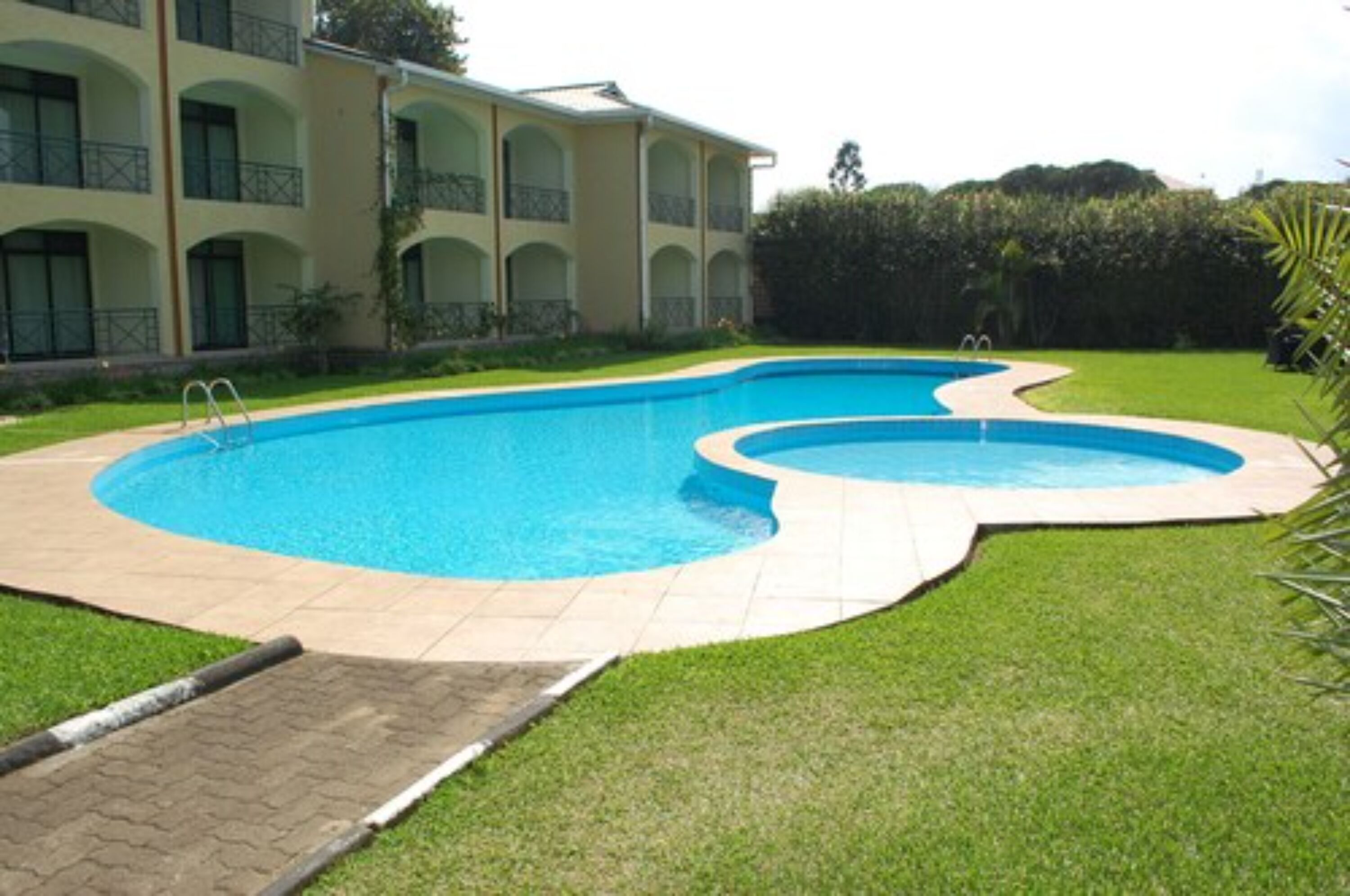 A swimming pool with a curved design, surrounded by a well-maintained lawn and a building with balconies.