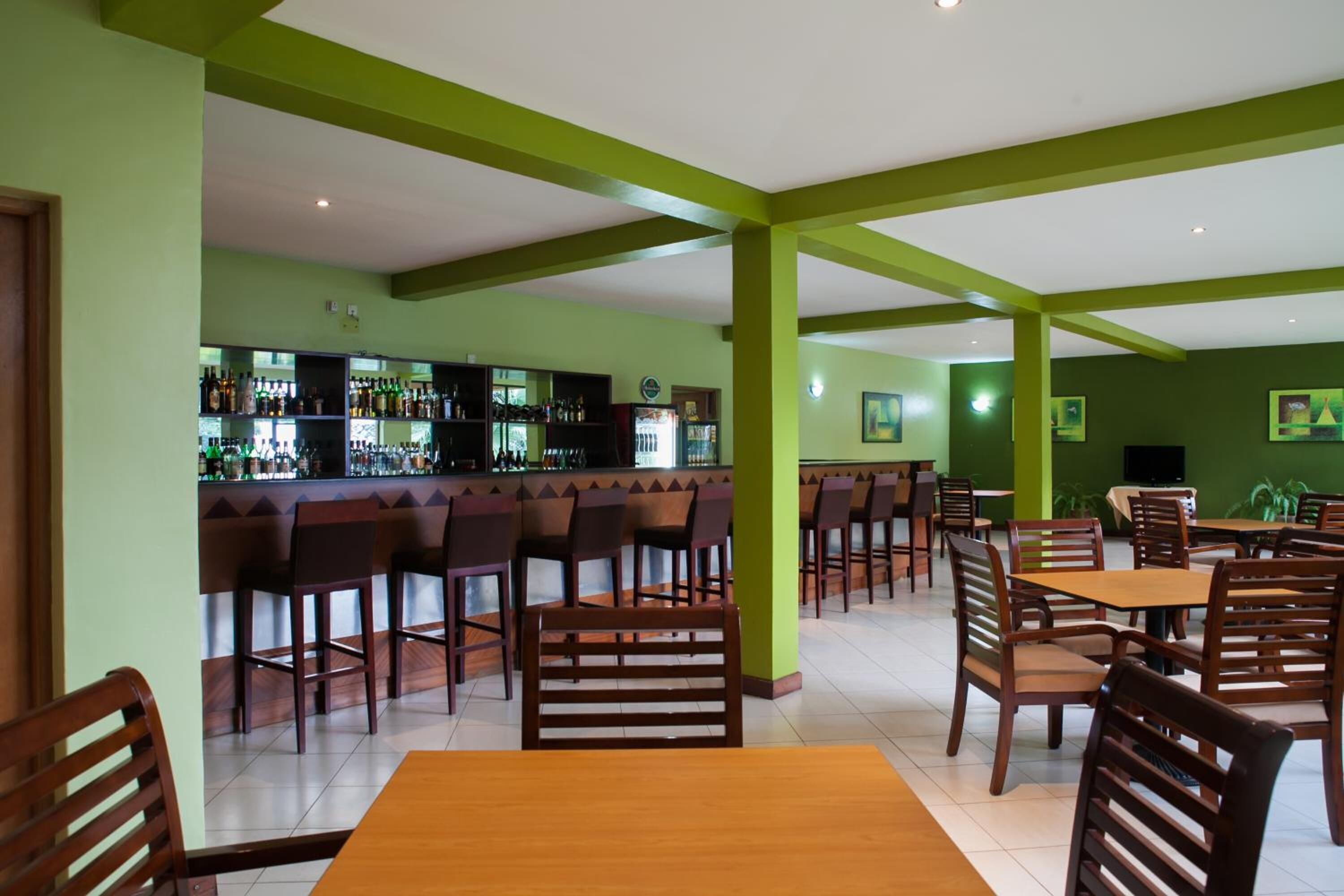 Um restaurante com paredes verdes, mesas e cadeiras de madeira, e um espaço de bar com prateleiras cheias de garrafas.