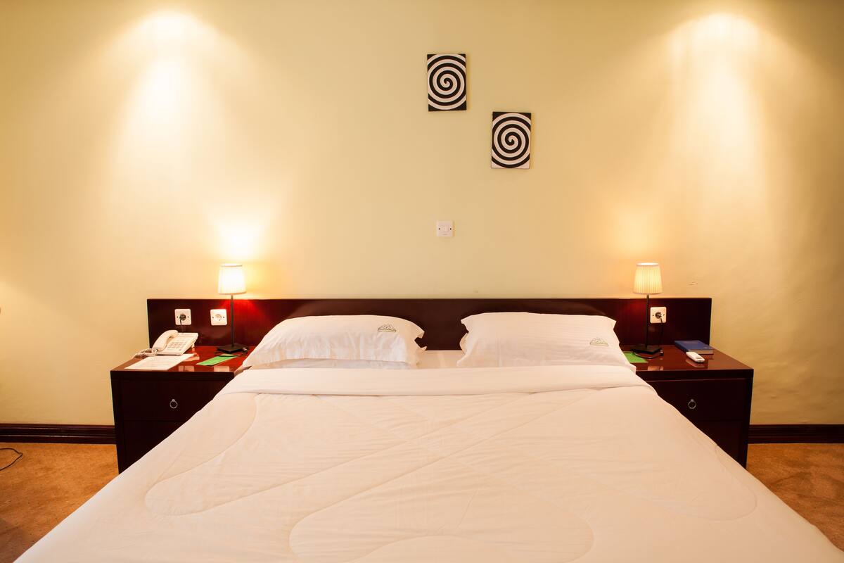 Habitación de hotel con una cama grande, dos mesitas de noche, un buró, una lámpara y dos luces fijadas en la pared.