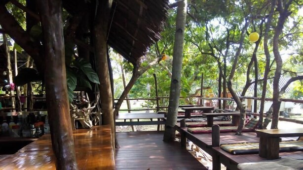 An outdoor wooden deck with benches and tables, surrounded by trees and a thatched roof structure. - Funky Fish Hostel (Ko Lanta)