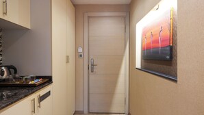 A hallway with a wooden door, a painting on the wall, and a kitchen area with a kettle and a tray.