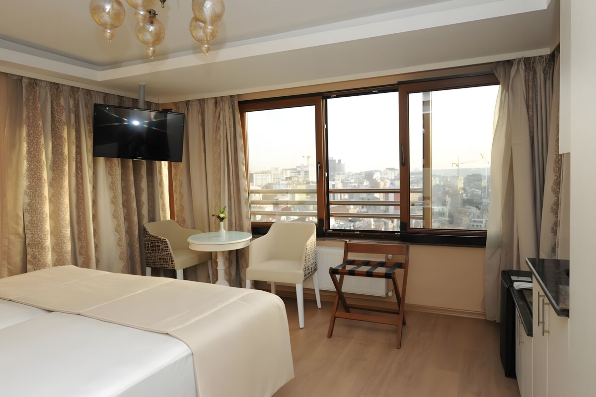 Une chambre d’hôtel équipée d’un lit, d’un bureau, d’une chaise, d’une télévision et d’une grande fenêtre donnant sur la ville.