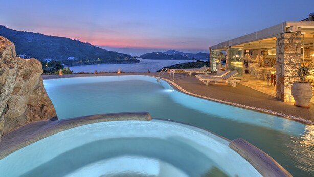 Zona de piscina con vista al mar y montañas, un edificio de piedra con fachada de vidrio y mobiliario exterior.