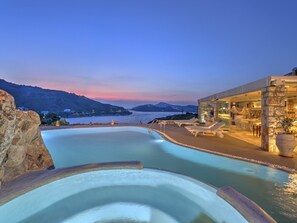 Zona piscina con vista sul mare e sulle montagne, edificio in pietra con facciata vetrata e arredi esterni.