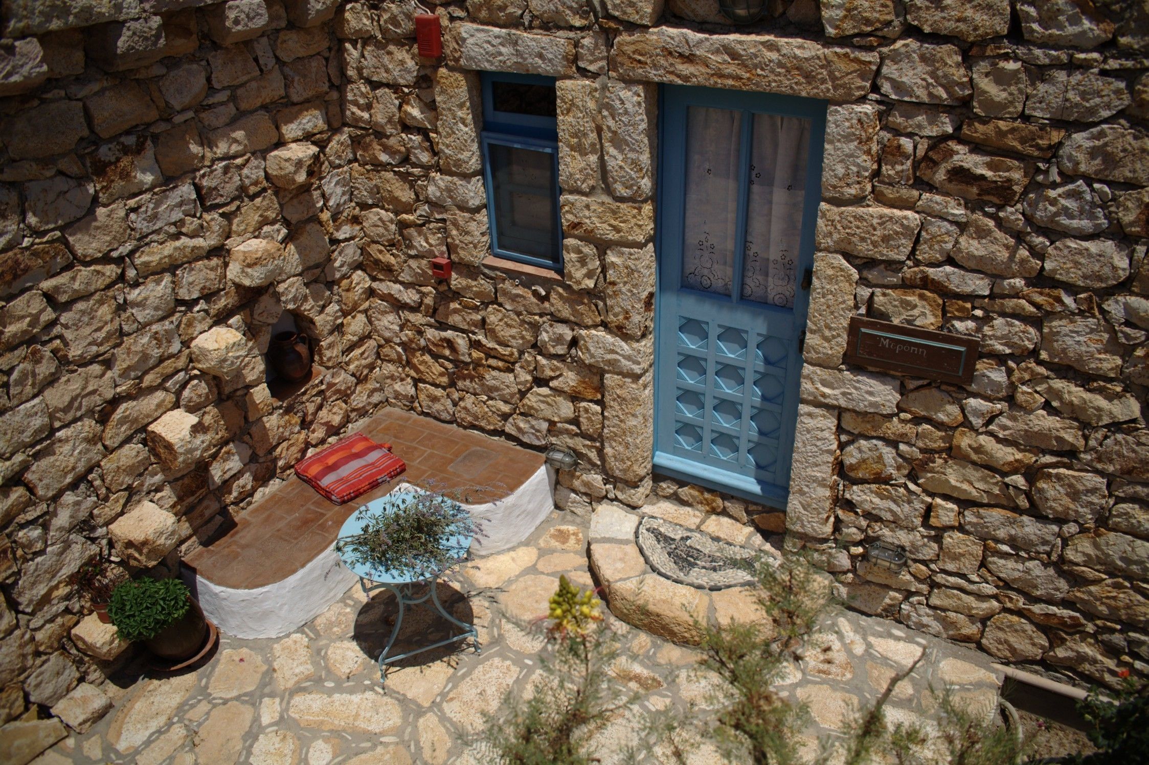 A stone building with a blue door and a small window. There is a red mat and a potted plant on the ground.