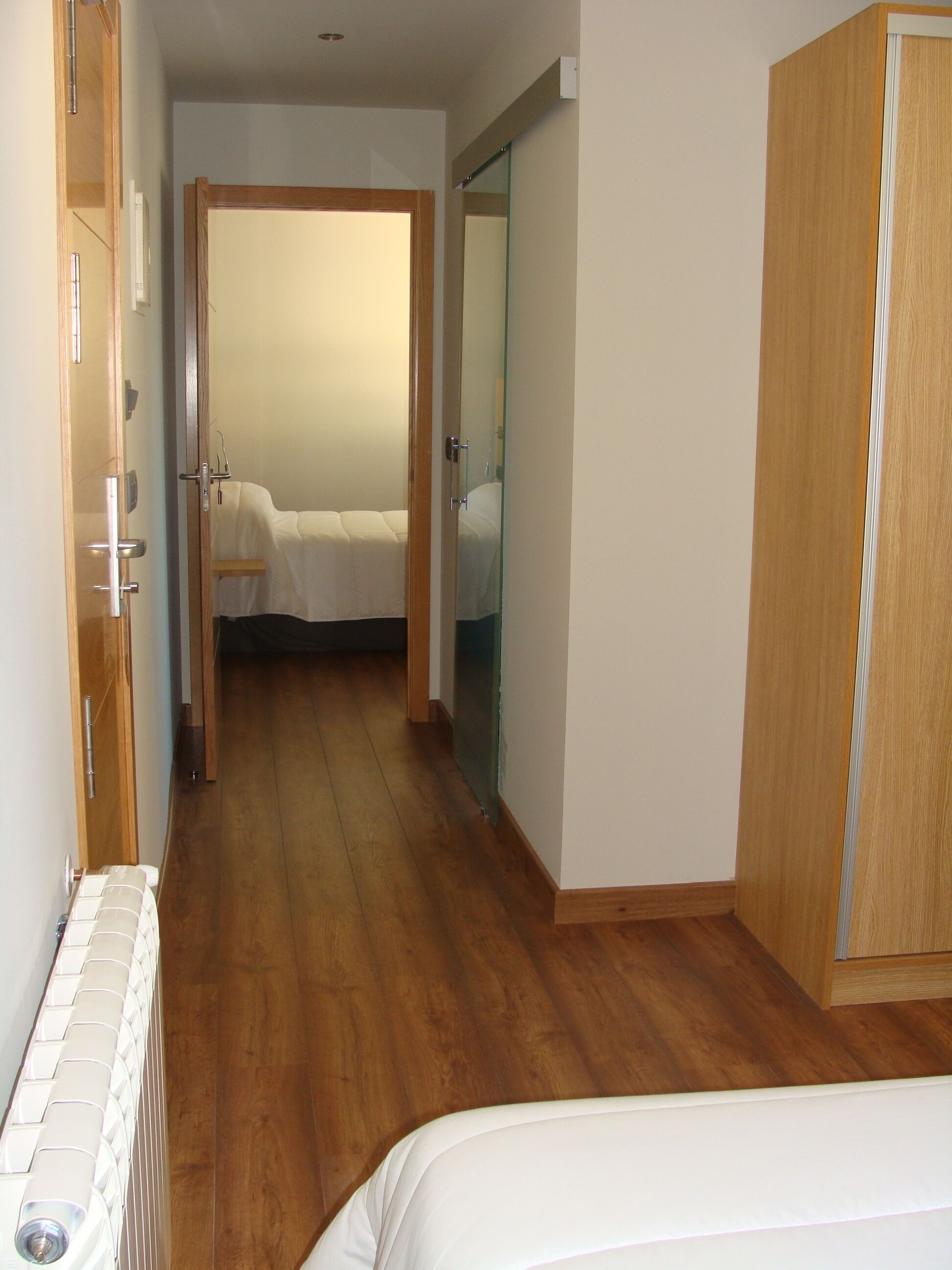 A narrow hallway with wooden flooring, a bed with white bedding in the distance, and a wardrobe on the right.