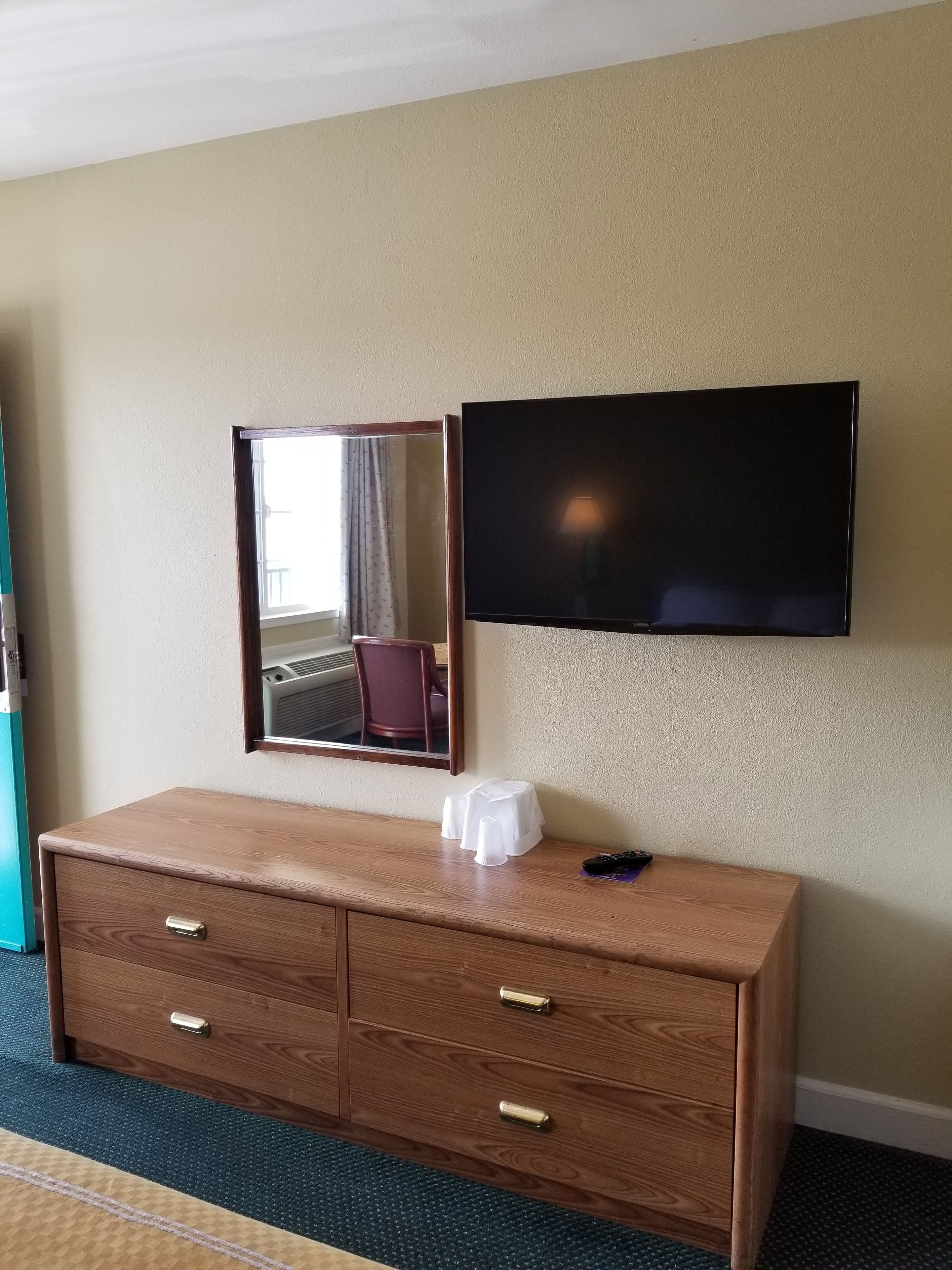 A hotel room with a wooden dresser, a flat-screen TV mounted on the wall, and a mirror above the dresser.