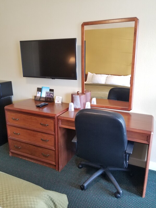 Une chambre d’hôtel avec un bureau en bois, un fauteuil de bureau noir, un téléviseur à écran plat, un miroir et un lit avec une literie blanche.