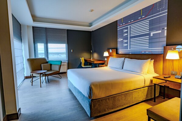 Habitación de hotel con una cama grande, un escritorio, una silla y vistas de la ciudad a través de la ventana.
