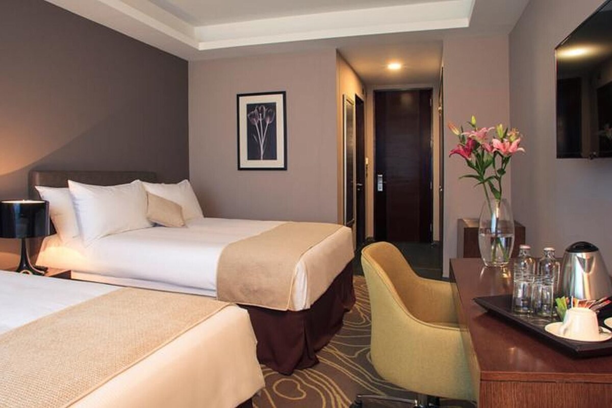 A hotel room with two beds, a desk with a chair, a vase with flowers, and a coffee maker.