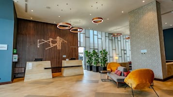 Zona de recepción de un hotel moderno con un mostrador de madera, un área de estar con sofá y sillones, y una lámpara decorativa.